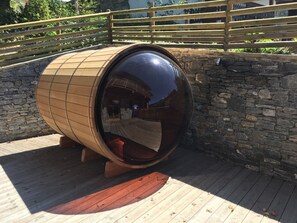 Uma estrutura em forma de barril de madeira com superfície refletora, sobre um deck de madeira e com um muro de pedra ao fundo.