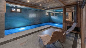 A spacious indoor swimming pool with a blue interior, wooden ceiling beams, and a seating area with two chairs.