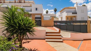 Un edificio residenziale dal design moderno, con una scala che conduce a un ingresso chiuso da cancello, circondato da un giardino curato con palme e fiori viola.