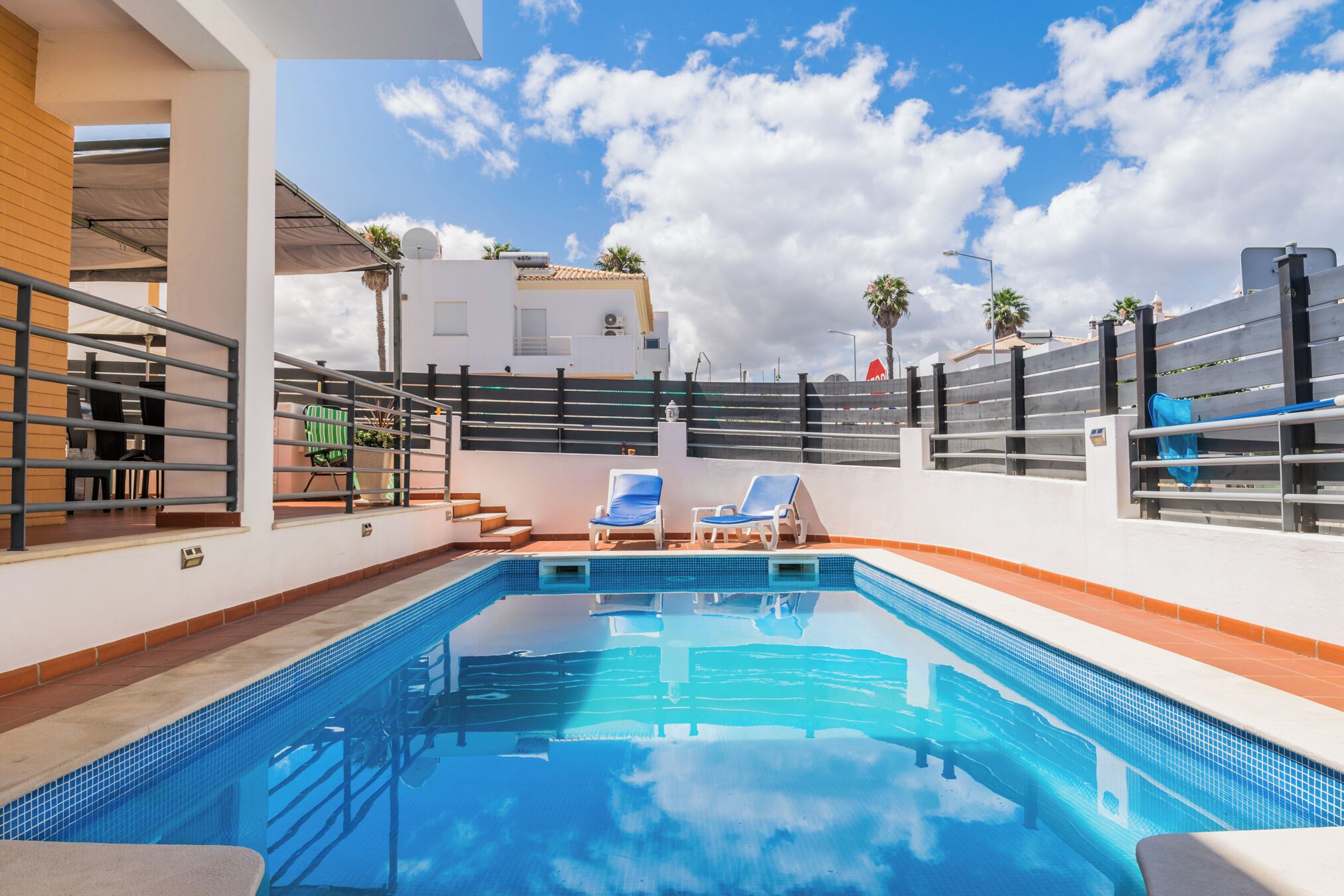 A swimming pool with two lounge chairs, a covered seating area, and a view of a white building with palm trees in the background.