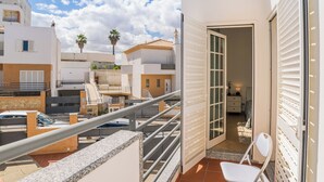 Un balcone con ringhiera in metallo, una sedia bianca e una porta che dà su una stanza interna con un letto e un comodino.