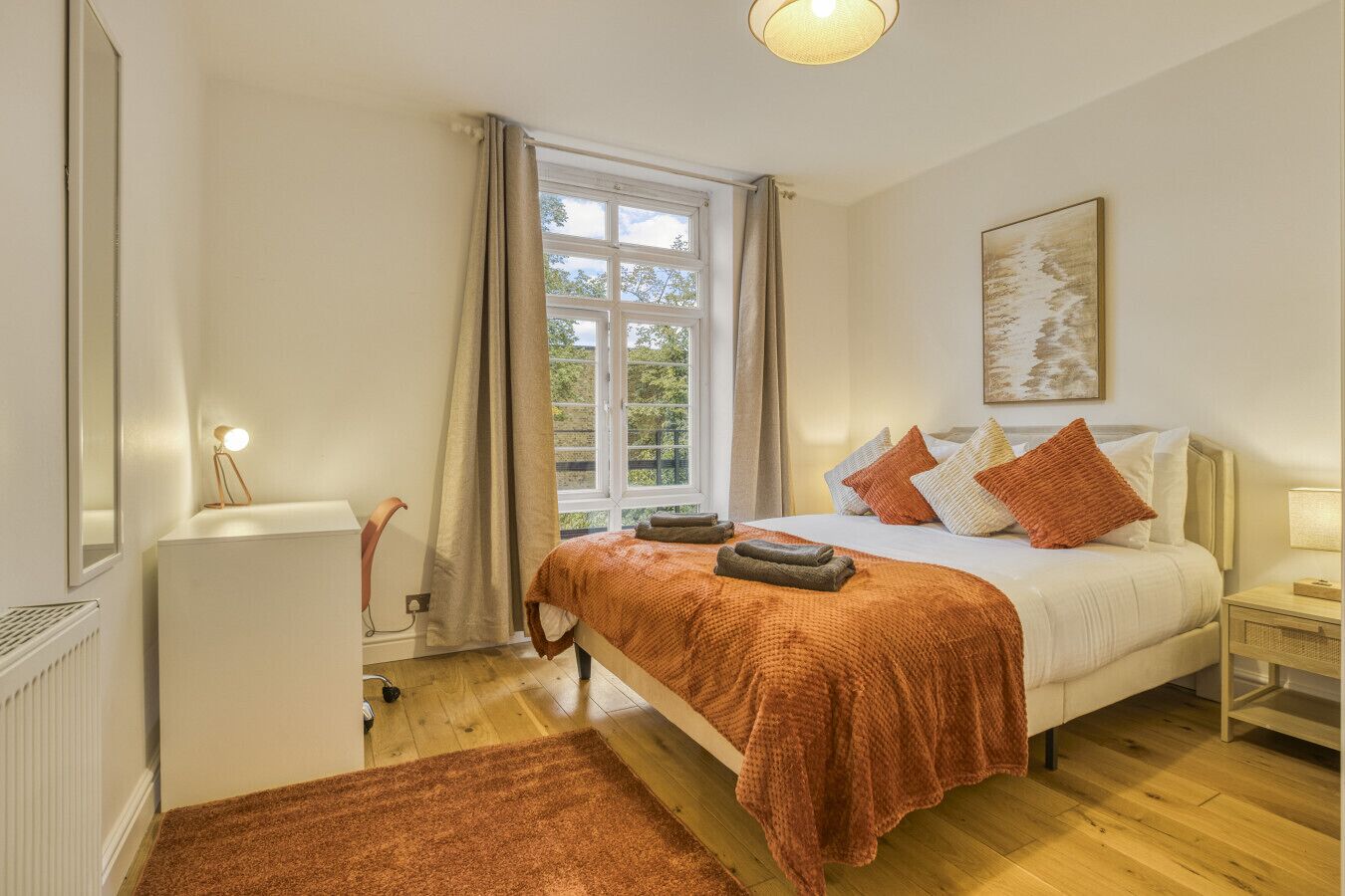 A bedroom with a bed, a nightstand, a lamp, a radiator, a window with curtains, and a painting on the wall.