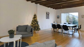 A modern living room with a wooden ceiling, a Christmas tree, a gray sofa, a dining table with chairs, and a view through a large window.