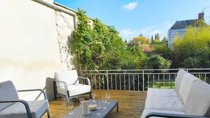 An outdoor patio with a wooden deck, two white cushioned chairs, a small table with a pitcher and wine glasses, and a white railing.