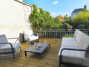 An outdoor patio with a wooden deck, two white cushioned chairs, a small table with a pitcher and wine glasses, and a white railing.