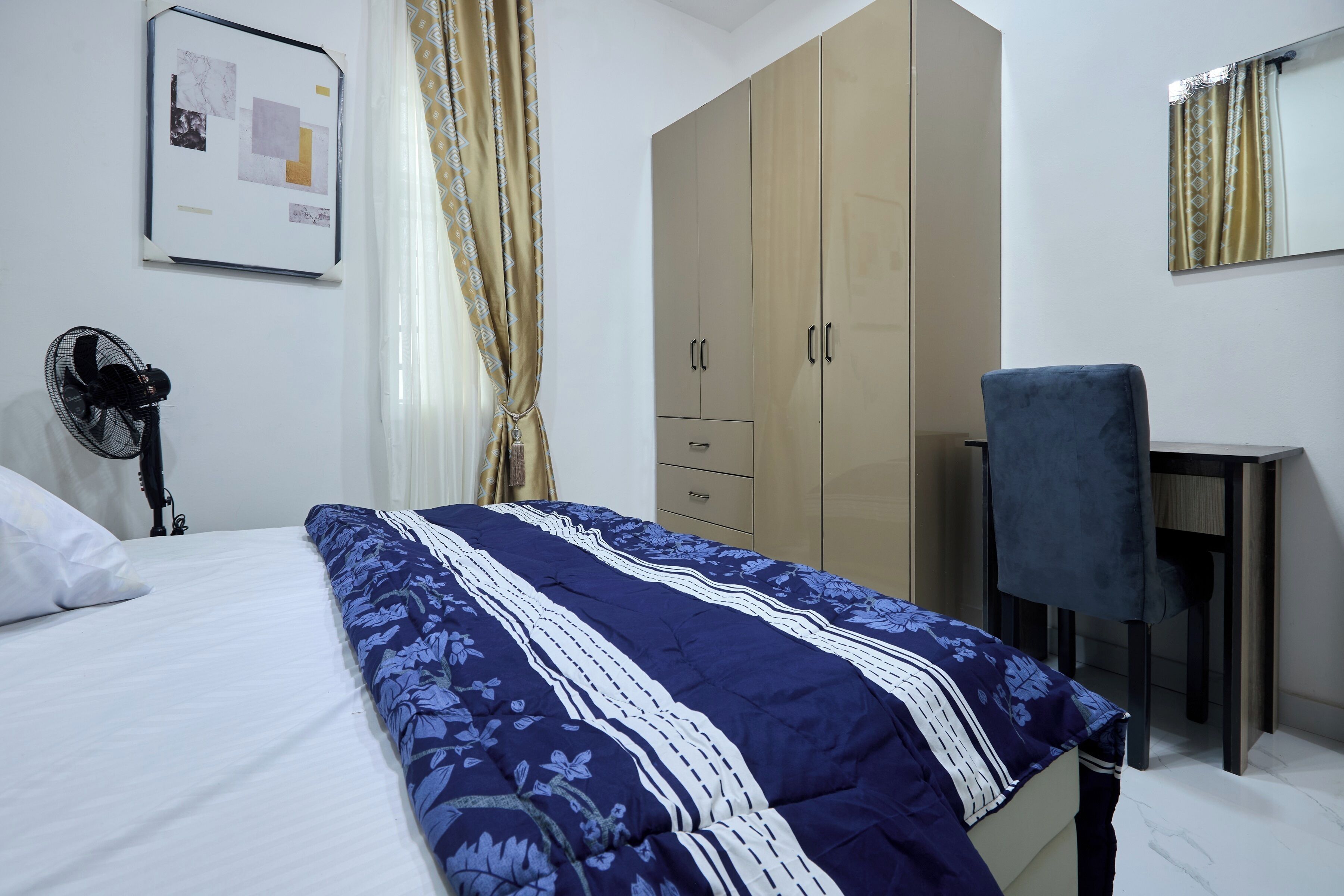 A bedroom with a bed, a desk, a chair, a wardrobe, and a fan.