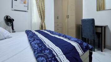 A bedroom with a bed, a desk, a chair, a wardrobe, and a fan.