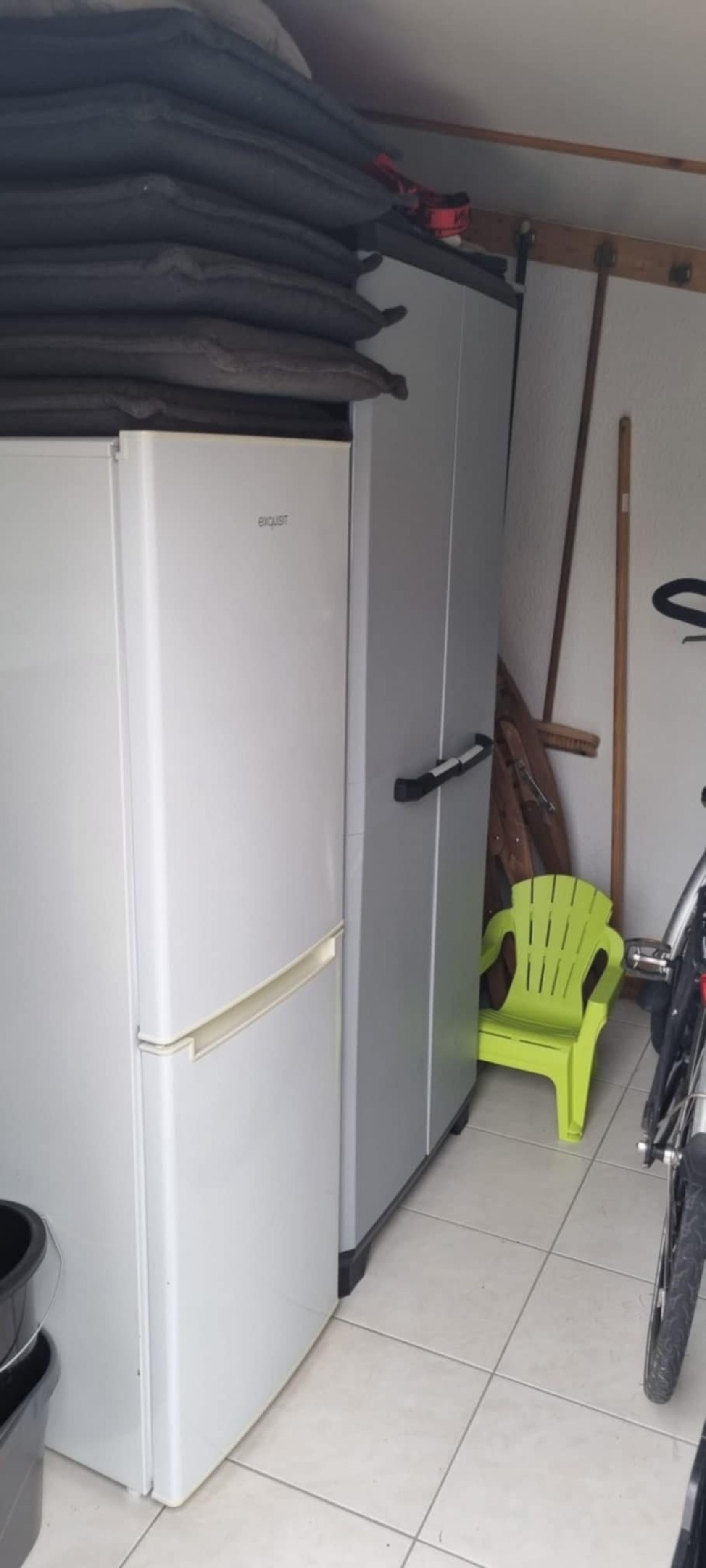 A compact kitchen with a refrigerator, a green plastic chair, and a broom leaning against the wall.
