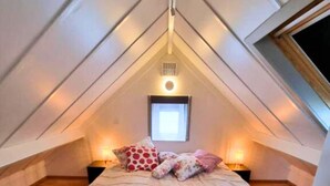 Un dormitorio con techo inclinado, una cama, dos mesitas de noche, un ventanal y un aplique de pared.