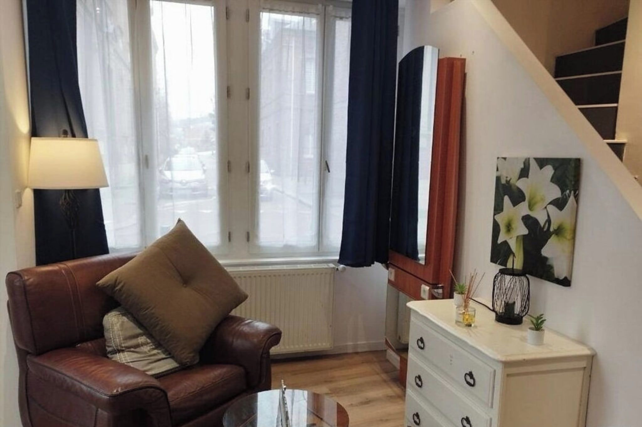 A living room with a brown leather sofa, a glass coffee table, and a white dresser with drawers. There is a window with curtains, a painting on the wall, and a staircase in the background.