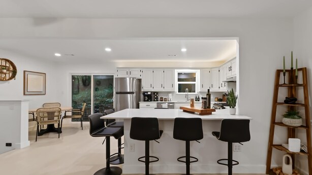 A modern kitchen with white cabinets, a central island with bar stools, and a dining area with wicker chairs. - War Eagle Haven, Lake Access, spacious deck, games (Springdale)