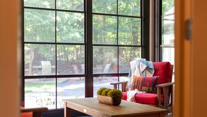 A cozy room with a large window, a red chair, a small table with a plant, and a colorful rug. - Tree Covered Cottage - Walk to Douglas Beach! (Douglas)