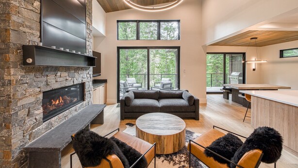 A modern living room with a fireplace, a sofa, a coffee table, and a kitchen area visible through large windows.