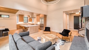 A modern kitchen with wooden cabinets and stainless steel appliances, a dining area with a grey sofa and a round coffee table, and a staircase with a stone wall.