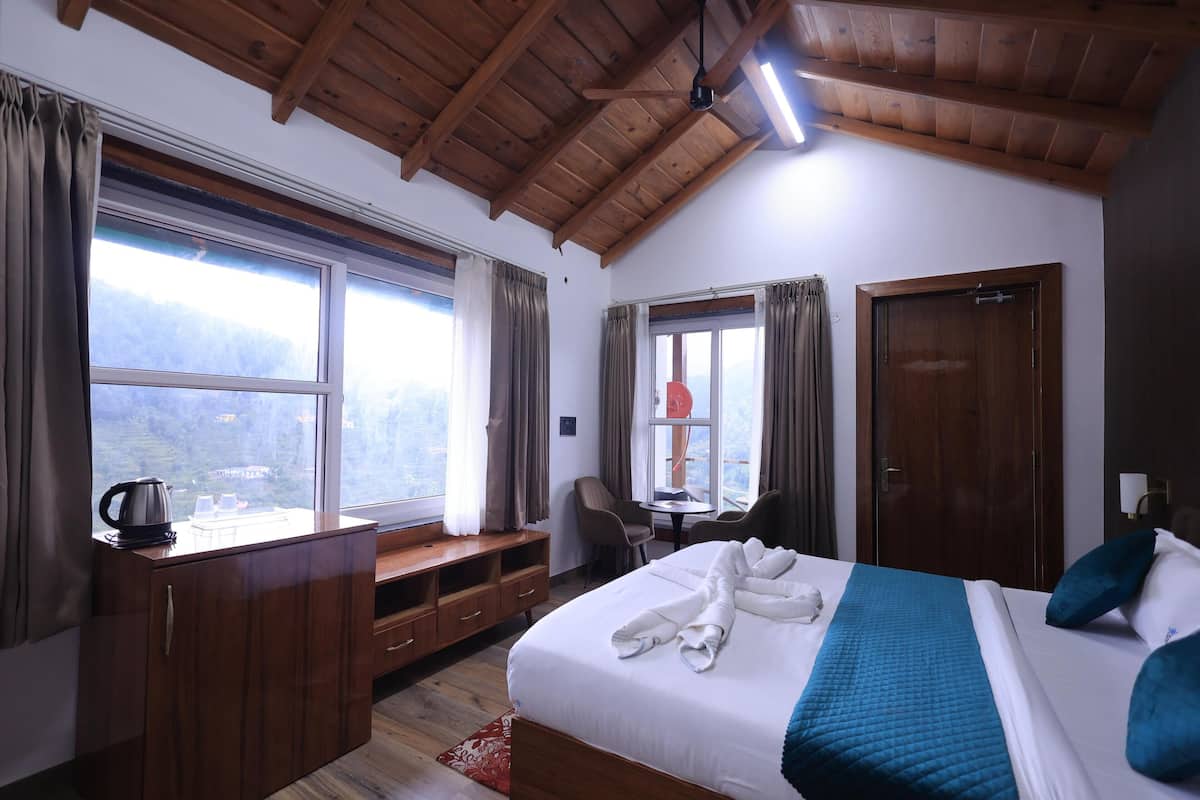 A hotel room with a large bed, wooden ceiling, a desk with a kettle, a chair, and a view of the mountains.