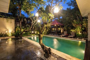Ein Pool mit Gazebo, umgeben von Palmen und Pflanzen, mit einer Terrasse und Stühlen.