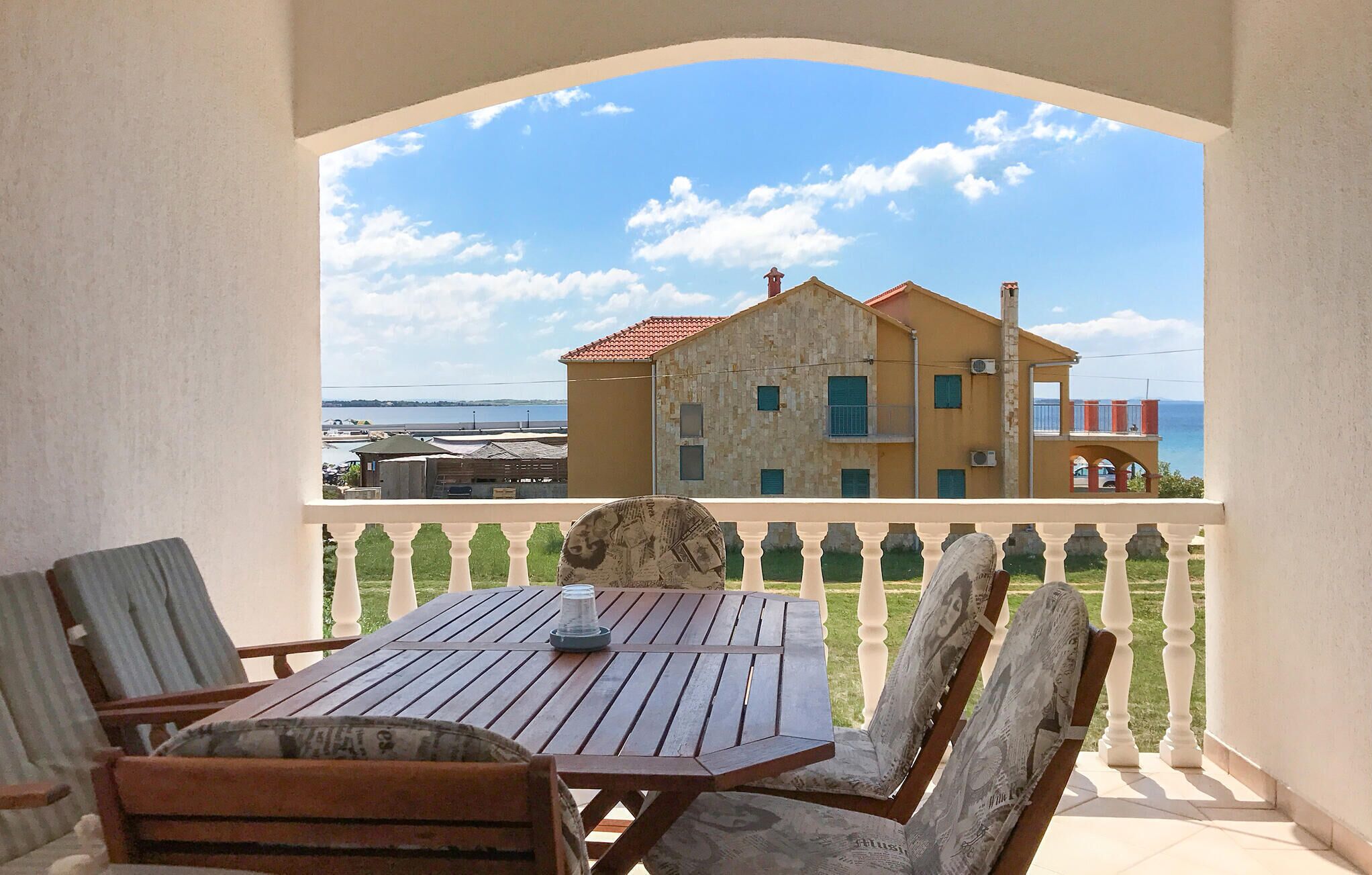 A balcony with a table and chairs, overlooking a building with a red roof and a view of the sea.