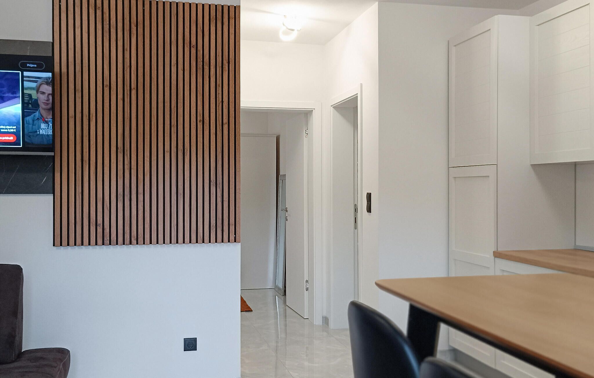 A modern kitchen with white cabinets, a wooden panel wall, and a television mounted on the wall.