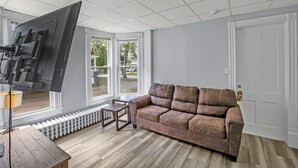 A living room with a brown leather sofa, a flat-screen TV mounted on the wall, a small wooden table, and a radiator under a window. - Division Champs 2 - All Star Baseball Rentals (Oneonta)
