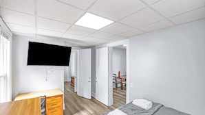 A hotel room with a bed, a desk with drawers, a television mounted on the wall, and an open door leading to another room. - Division Champs 2 - All Star Baseball Rentals (Oneonta)