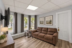 A living room with a brown sofa, a small table, a lamp, a radiator, and a TV mounted on the wall. - Division Champs 2 - All Star Baseball Rentals (Oneonta)