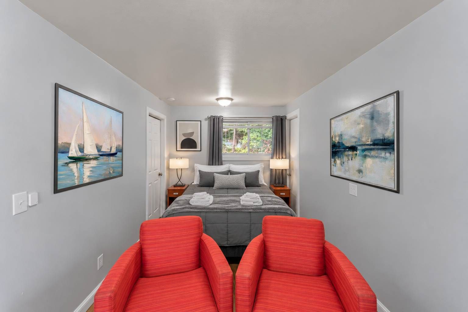 A hotel room with a bed, two red chairs, a window with curtains, and two paintings on the walls.