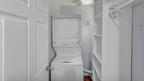 A compact laundry room with a built-in washer and dryer, a ceiling vent, and a metal shelf unit. - Main Lineup Apt 3 - All Star Baseball Rentals (Oneonta)