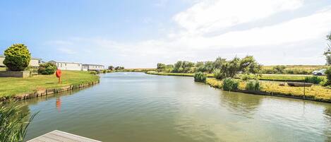 A calm river with a wooden dock, grassy banks, and a row of houses in the background.