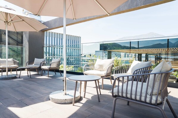 Une terrasse sur le toit avec des sièges extérieurs, une balustrade en verre et une vue sur un immeuble moderne et des montagnes.