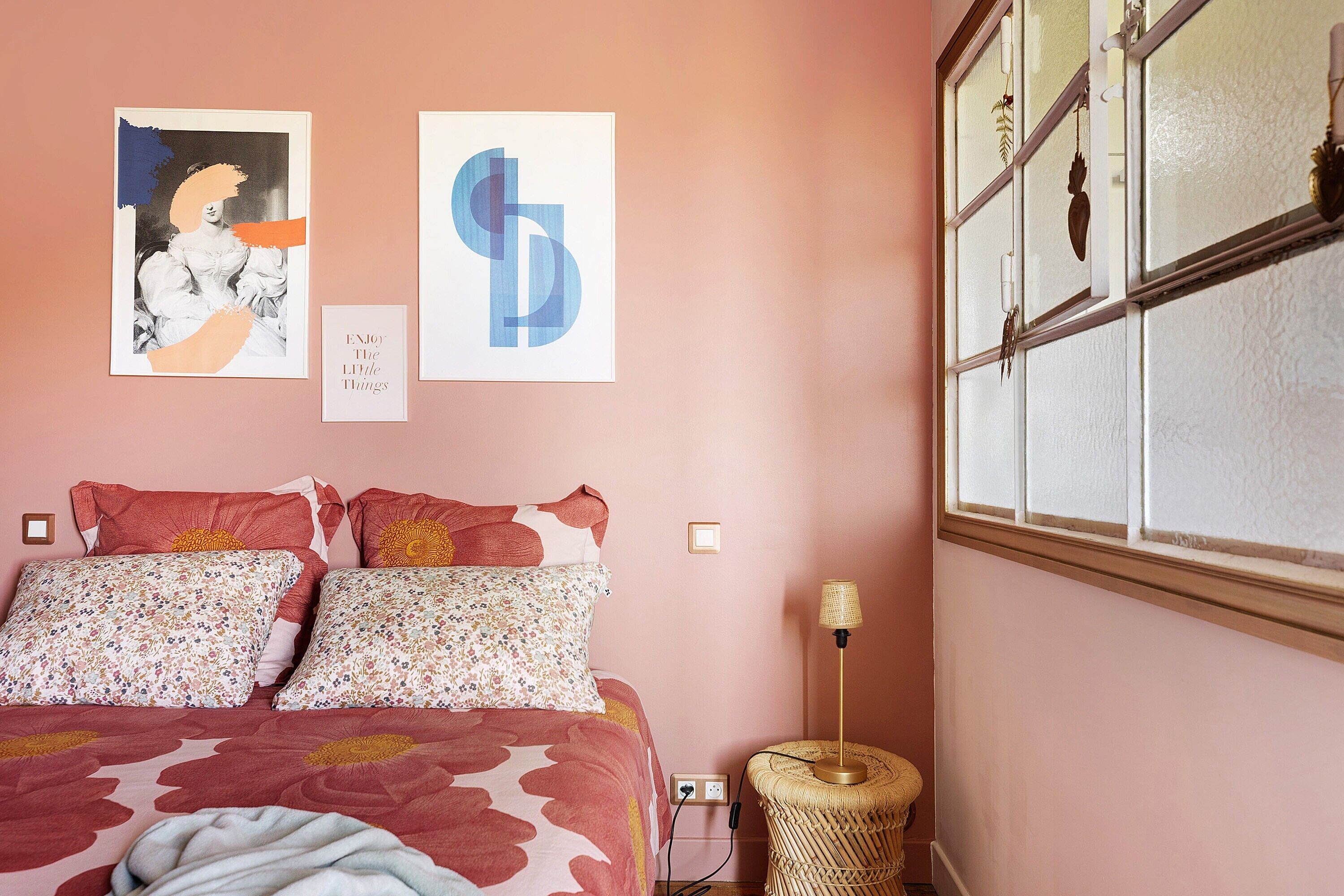 A bedroom with a bed, floral pillows, a wall with artwork, a window, and a bedside lamp.