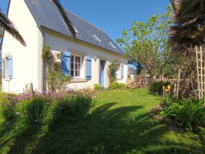 Ein Haus mit geneigtem Dach, blauen Fensterläden und einem Garten mit verschiedenen Pflanzen und Blumen.