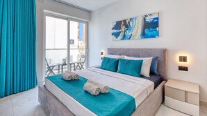 A modern hotel room with a large bed, a balcony with chairs, and a city view.