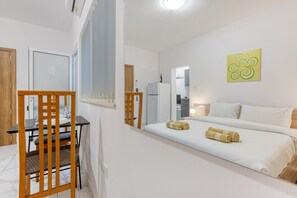 Une chambre à coucher avec un lit, une chaise en bois, une table et un coin cuisine visible à travers une porte ouverte.