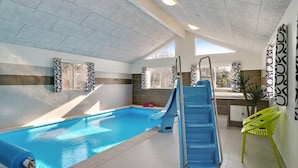 Une piscine intérieure avec un toboggan bleu, des murs carrelés et une vue sur les arbres à l’extérieur.
