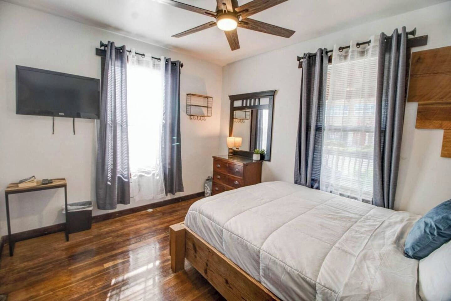 A bedroom with a bed, a dresser, a TV mounted on the wall, and a ceiling fan.