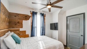 A bedroom with a bed, wooden headboard, curtains, a ceiling fan, a television, and a door.
