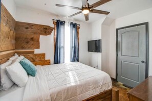 A bedroom with a bed, wooden headboard, curtains, a ceiling fan, a television, and a door.