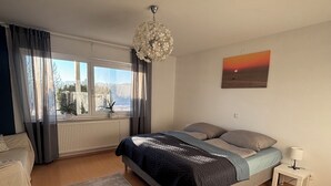 Camera da letto con un letto, un comodino, una finestra con tende, un termosifone, una lampada a sospensione e un quadro appeso al muro.