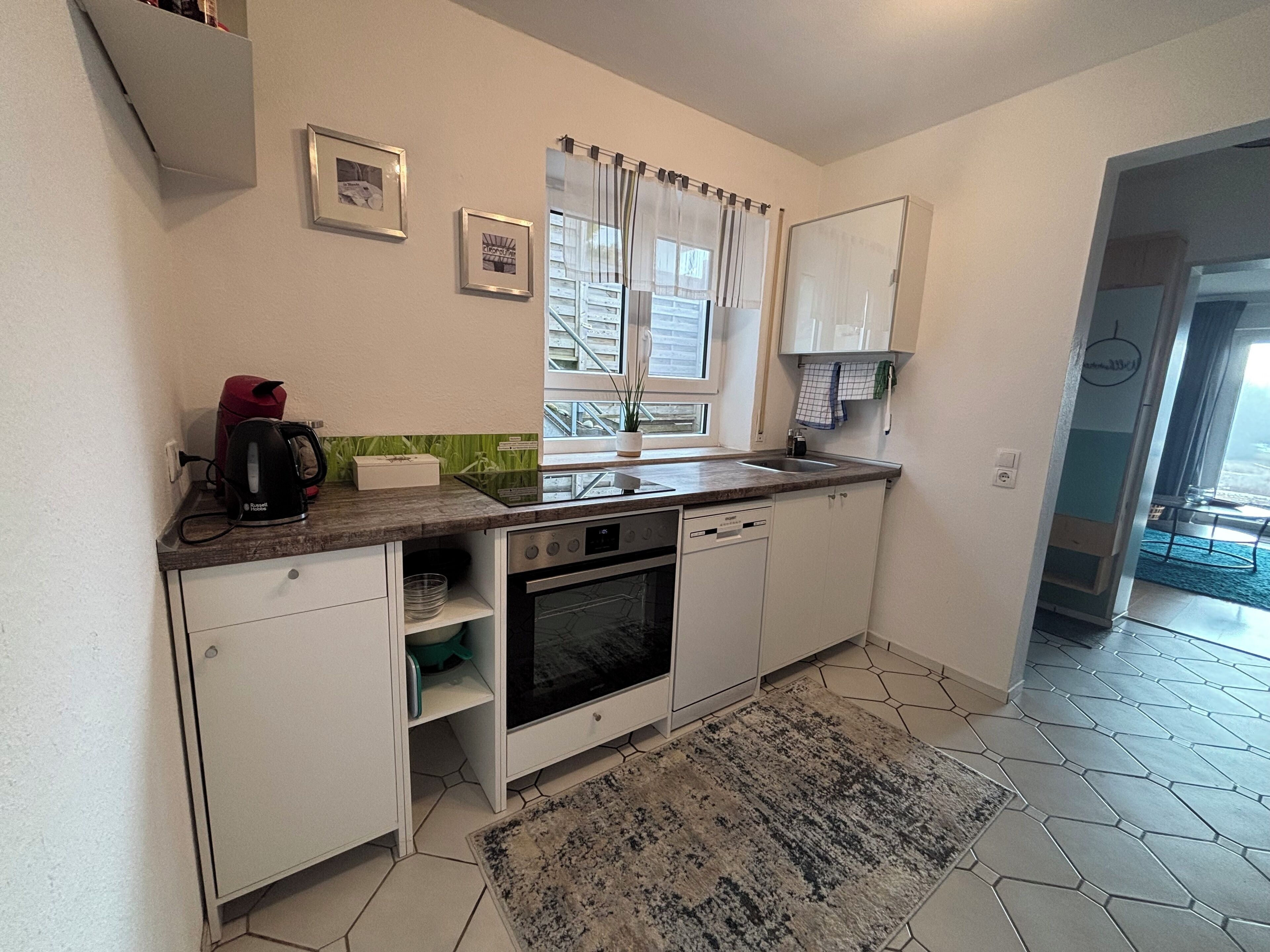 Una cucina compatta con mobili bianchi, forno integrato, lavastoviglie e una finestra con tende.