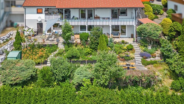 Una casa a due piani con tetto in tegole rosse, un balcone e un giardino con diverse piante e un piccolo stagno.