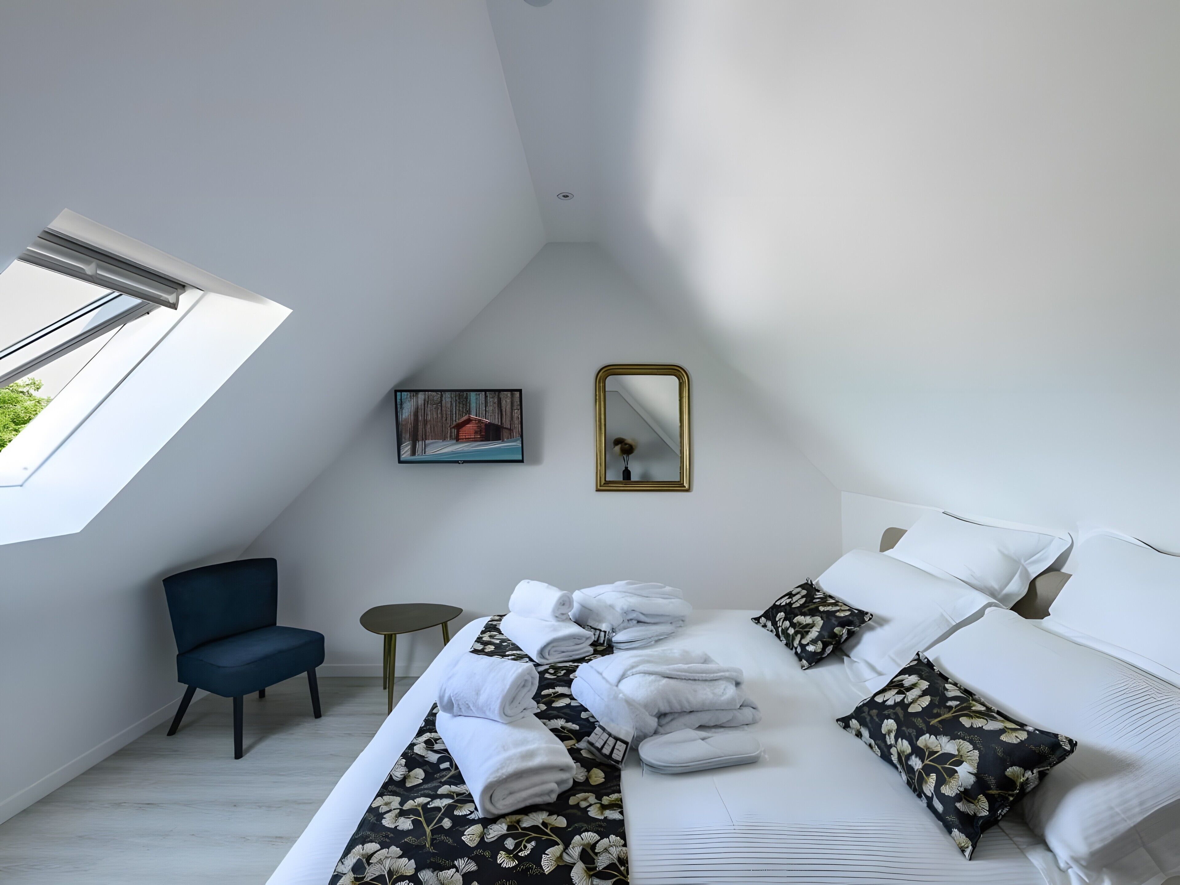 Une chambre avec une lucarne, un lit avec des serviettes et des chaussons, une chaise, une petite table et un tableau au mur.