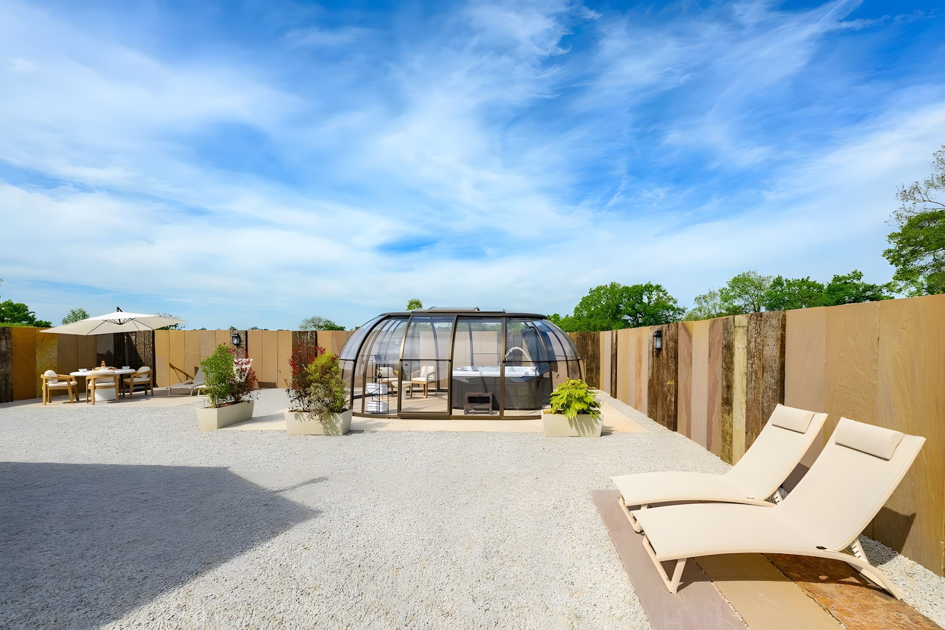 Une serre à structure transparente, dotée d’un espace repas et de fauteuils pour se détendre à l’extérieur.