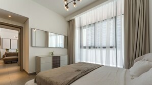 A modern bedroom with a large bed, a bedside table, a mirror, and a window with curtains. - Modern 1 BR Oxford Terraces (Dubai)