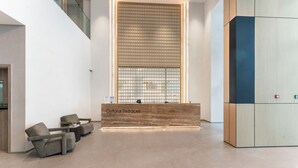 A reception area with a wooden reception desk labeled "Oxford Terraces," two grey armchairs, and a large vertical screen. - Modern 1 BR Oxford Terraces (Dubai)