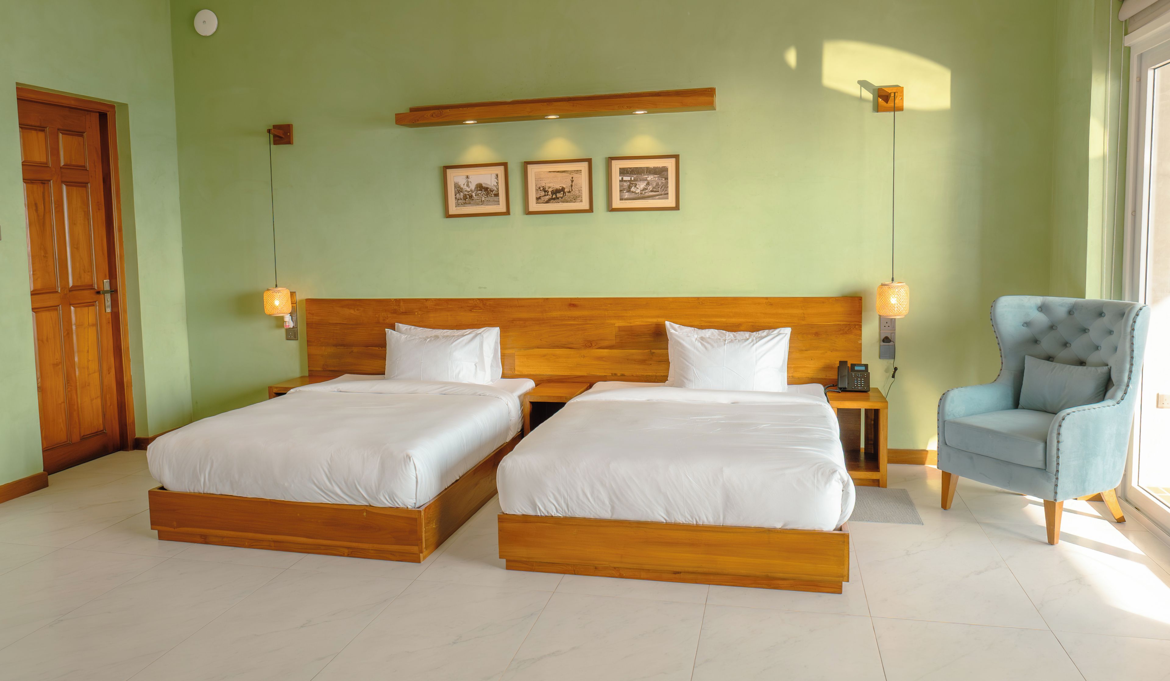 Une chambre d’hôtel avec deux lits en bois, une chaise, une table de chevet avec un téléphone et des tableaux accrochés au mur.