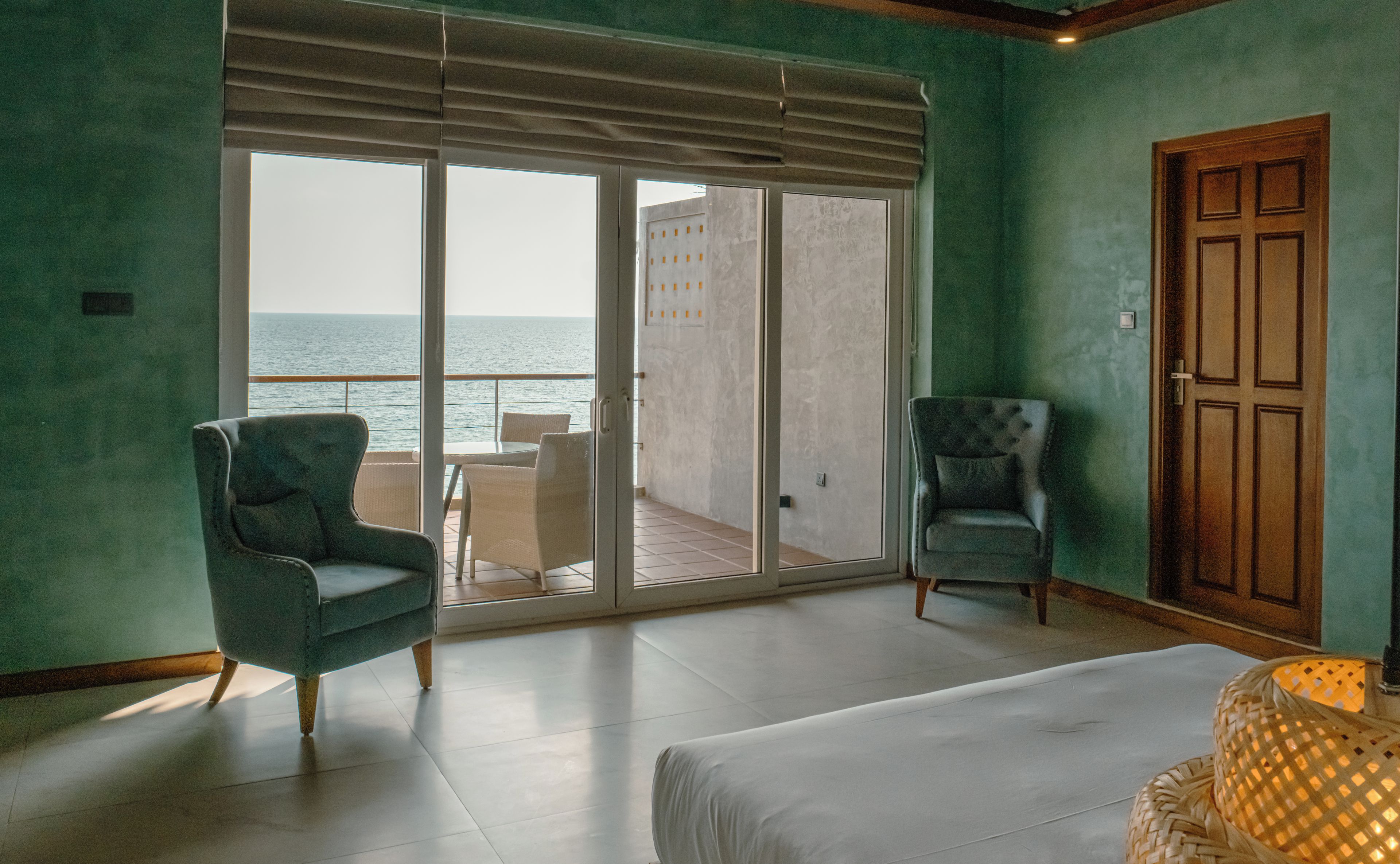 A room with a large sliding glass door leading to a balcony with a view of the sea. There are two chairs and a bed in the room.