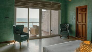 A room with a large sliding glass door leading to a balcony with a view of the sea. There are two chairs and a bed in the room.