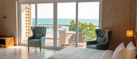 Une chambre d’hôtel avec un grand lit, deux fauteuils, une vue sur la mer et un balcon aménagé avec du mobilier d’extérieur.
