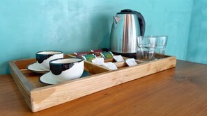 Una bandeja de madera con dos tazas, una tetera, bolsitas de té y vasos sobre una mesa de madera frente a una pared verde azulado.