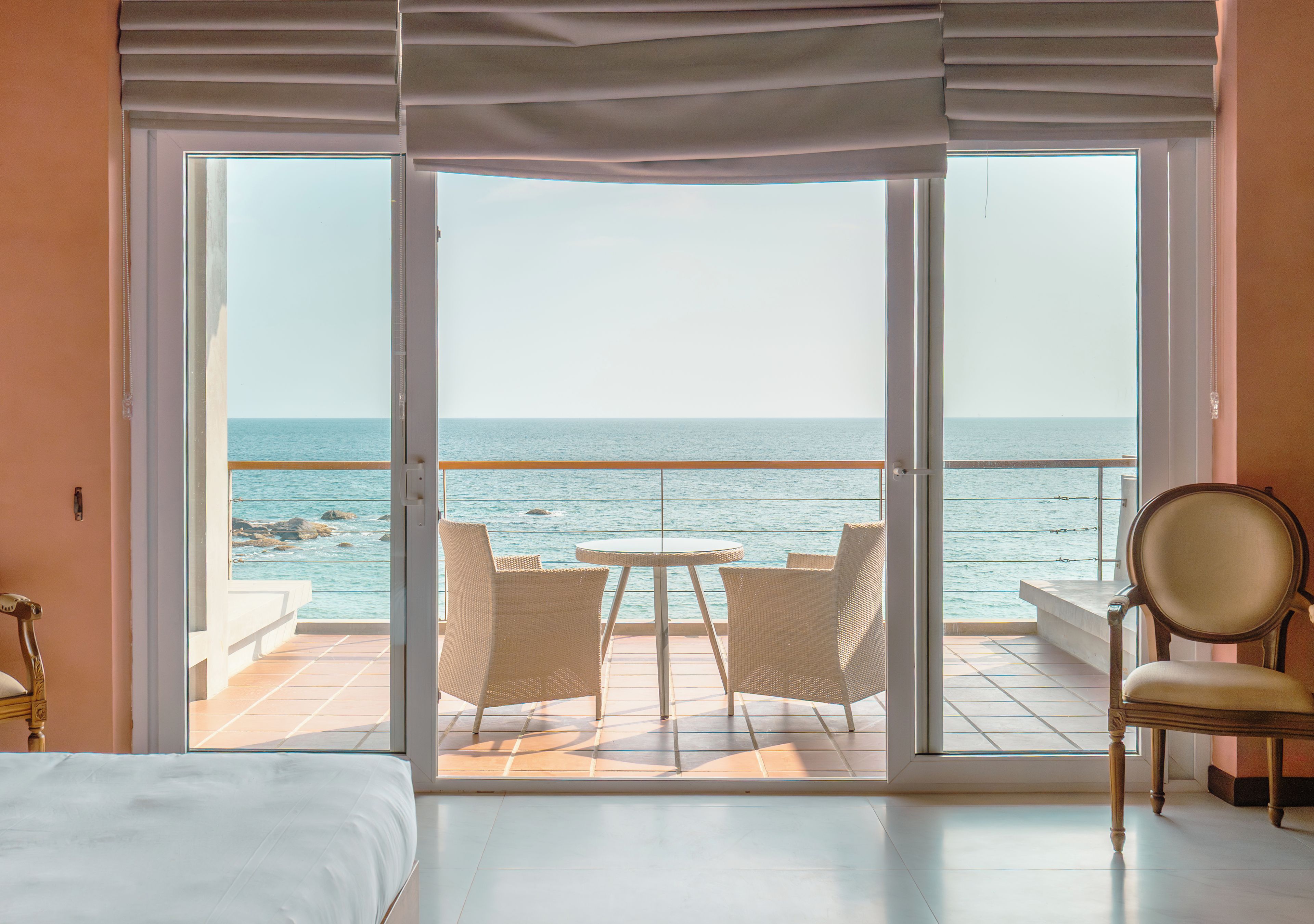 A hotel room with a large sliding glass door leading to a balcony with a view of the sea. There is a bed, a chair, and a small table on the balcony.